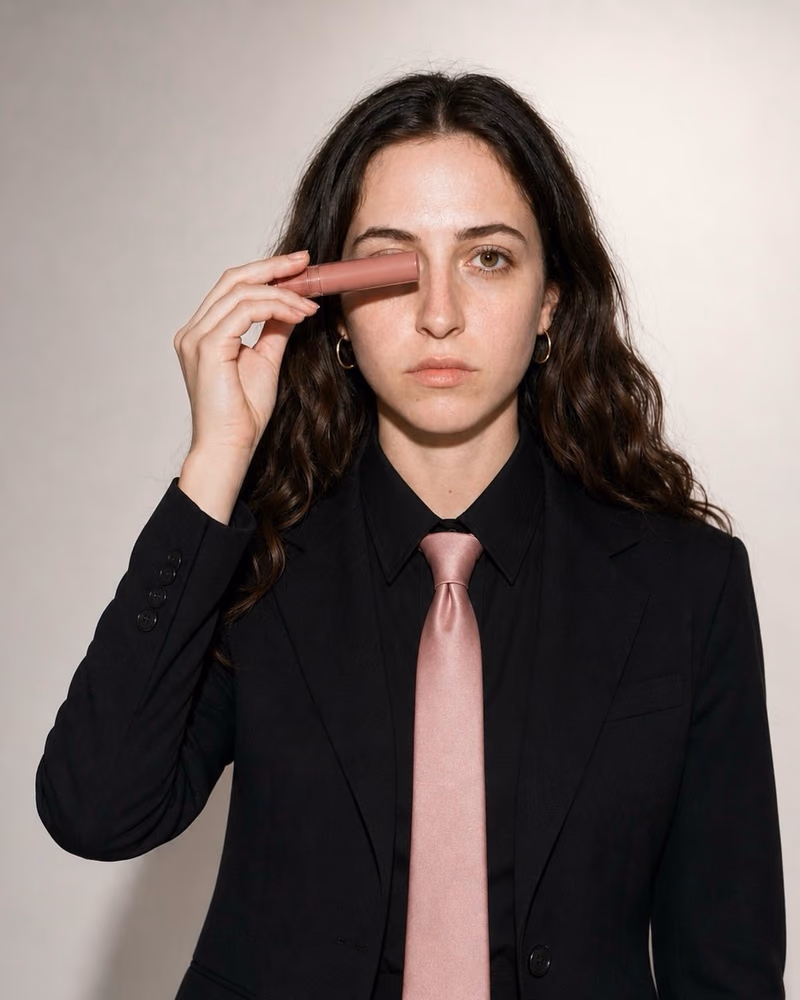 Hson campaign portrait: model in a black suit and pink tie holding a lipstick to one eye
